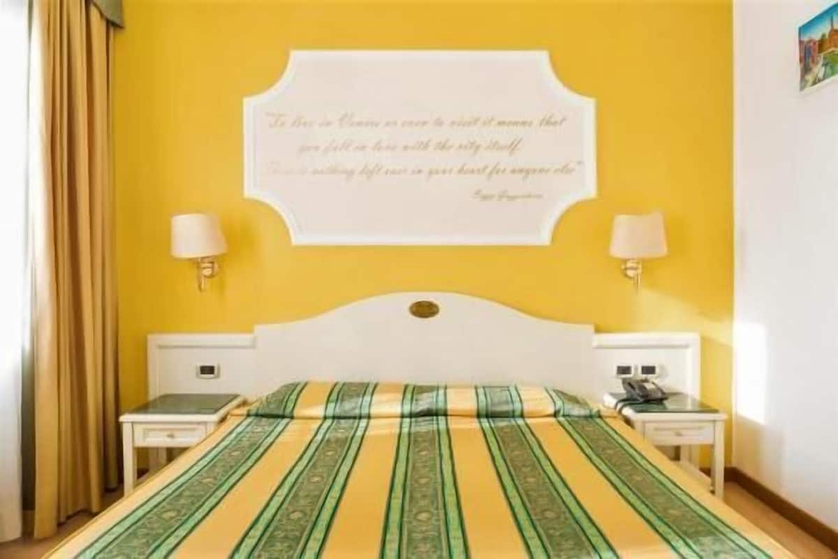 A hotel room with a yellow wall, a decorative quote, two bedside lamps, a bed with a striped bedspread, and a nightstand with a telephone.