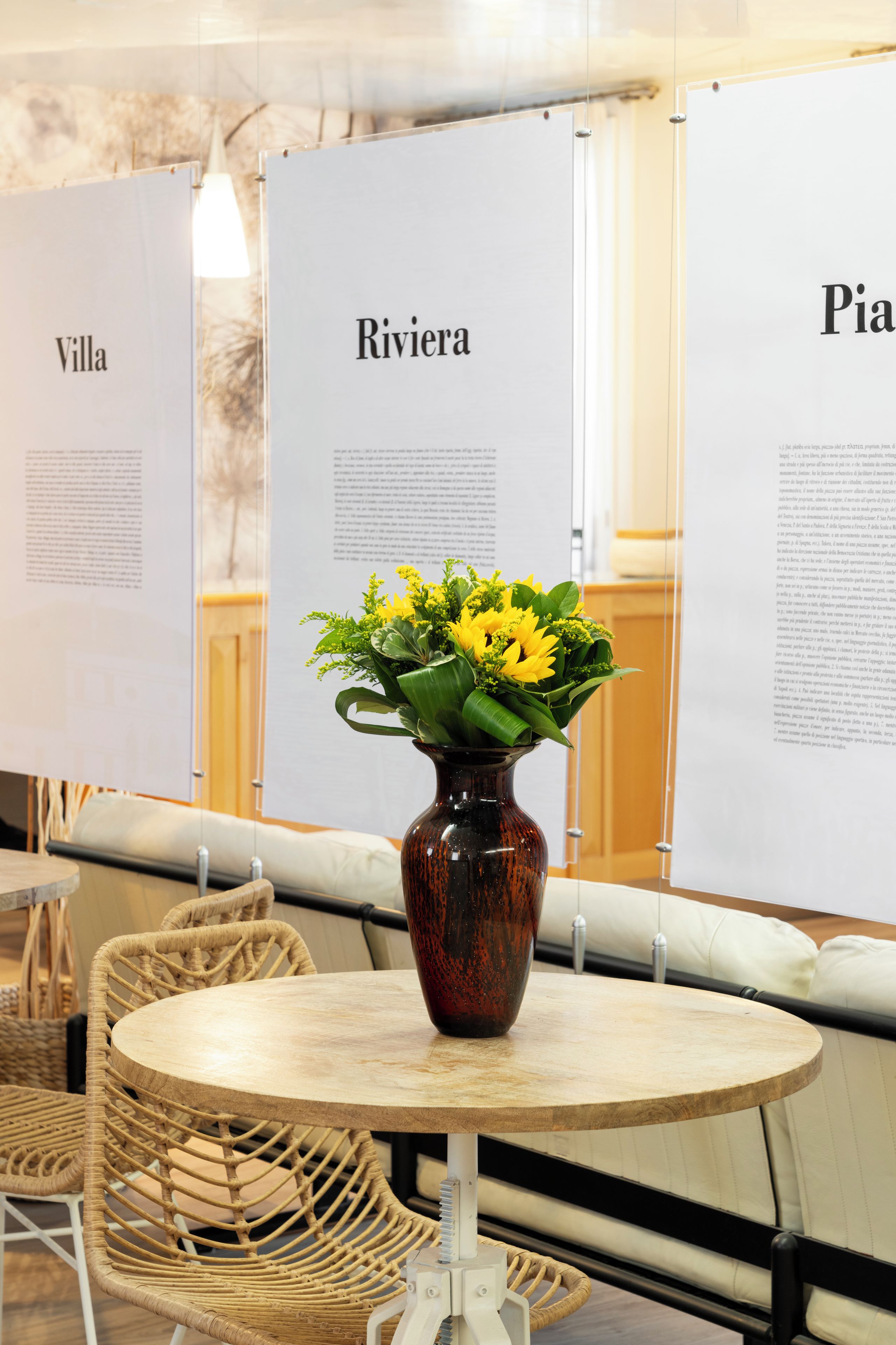 A round table with a dark brown vase containing yellow flowers, set against a backdrop of white panels with text.