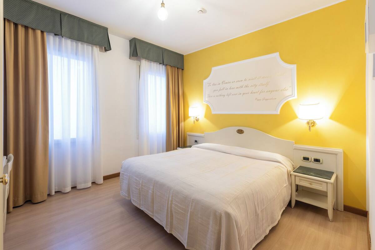 A hotel room with a bed, curtains, a wall quote, and bedside lamps.