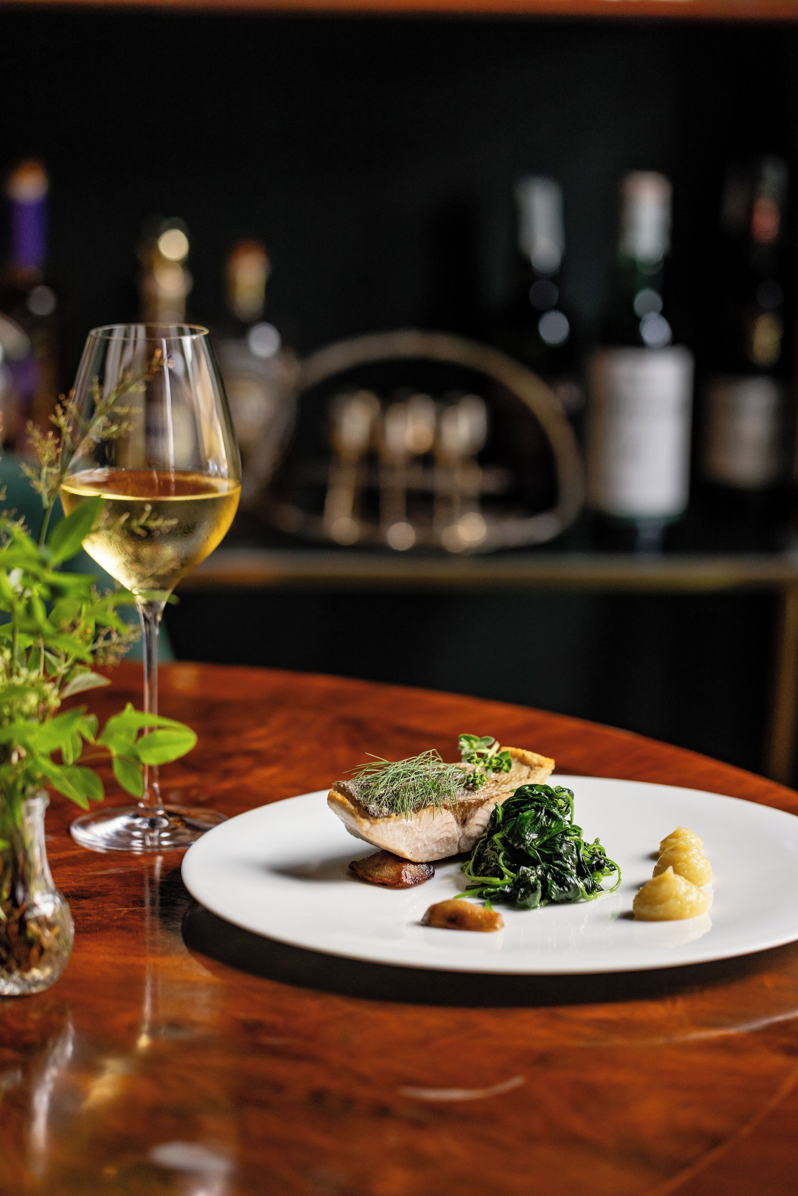 A dining table with a plate of food, a glass of white wine, and a vase of greenery.