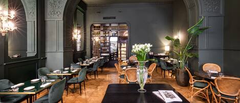 A restaurant interior with a central black table, green upholstered chairs, and a bar area in the background.