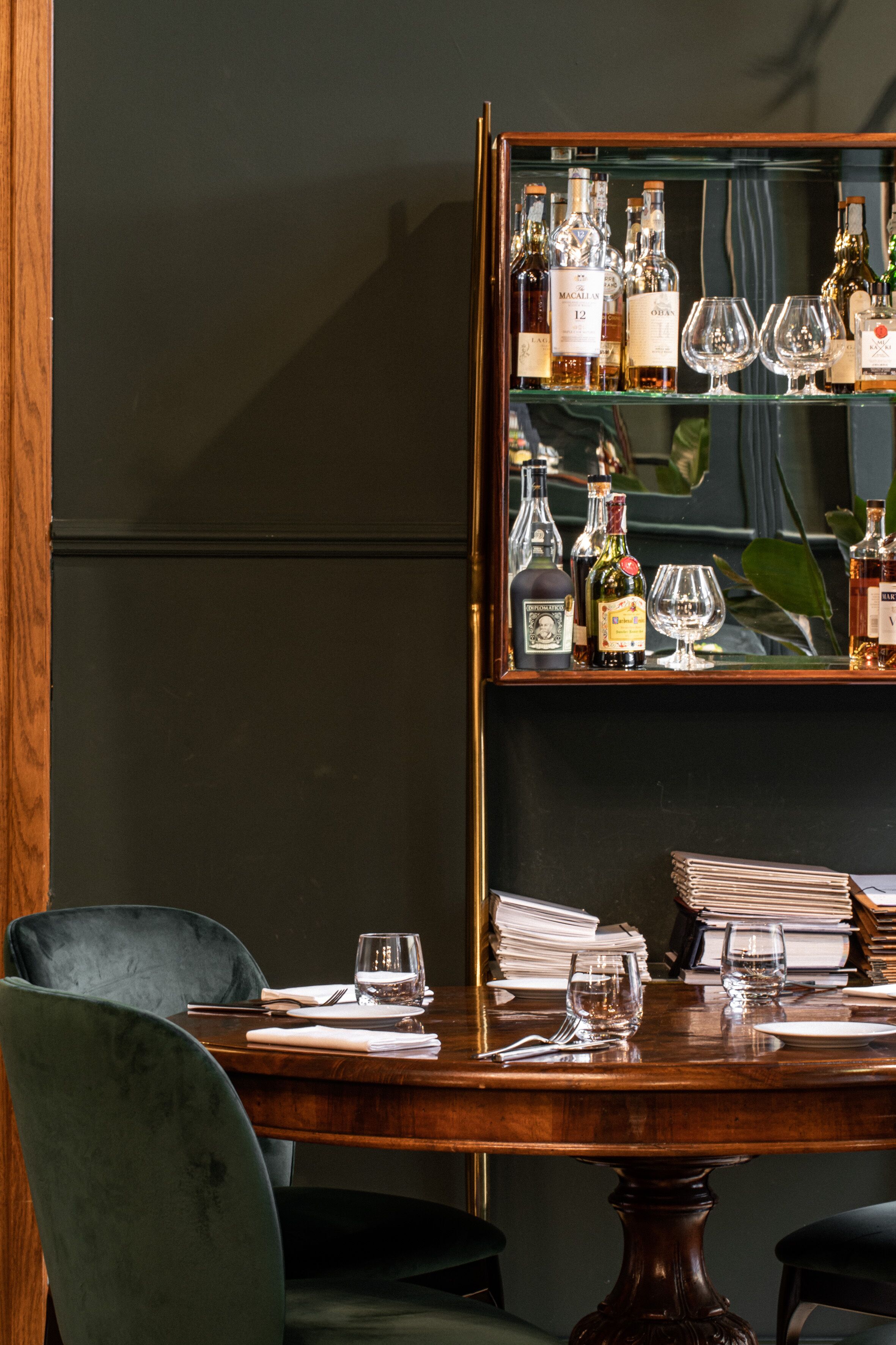 Un comedor con una mesa de madera dispuesta para dos personas, un espejo con un expositor de licores y sillas tapizadas en color verde oscuro.