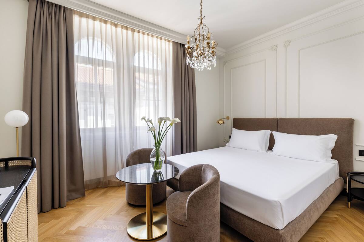 Una habitación de hotel con una cama grande, una mesa redonda con sillas, un jarrón con flores y una araña.