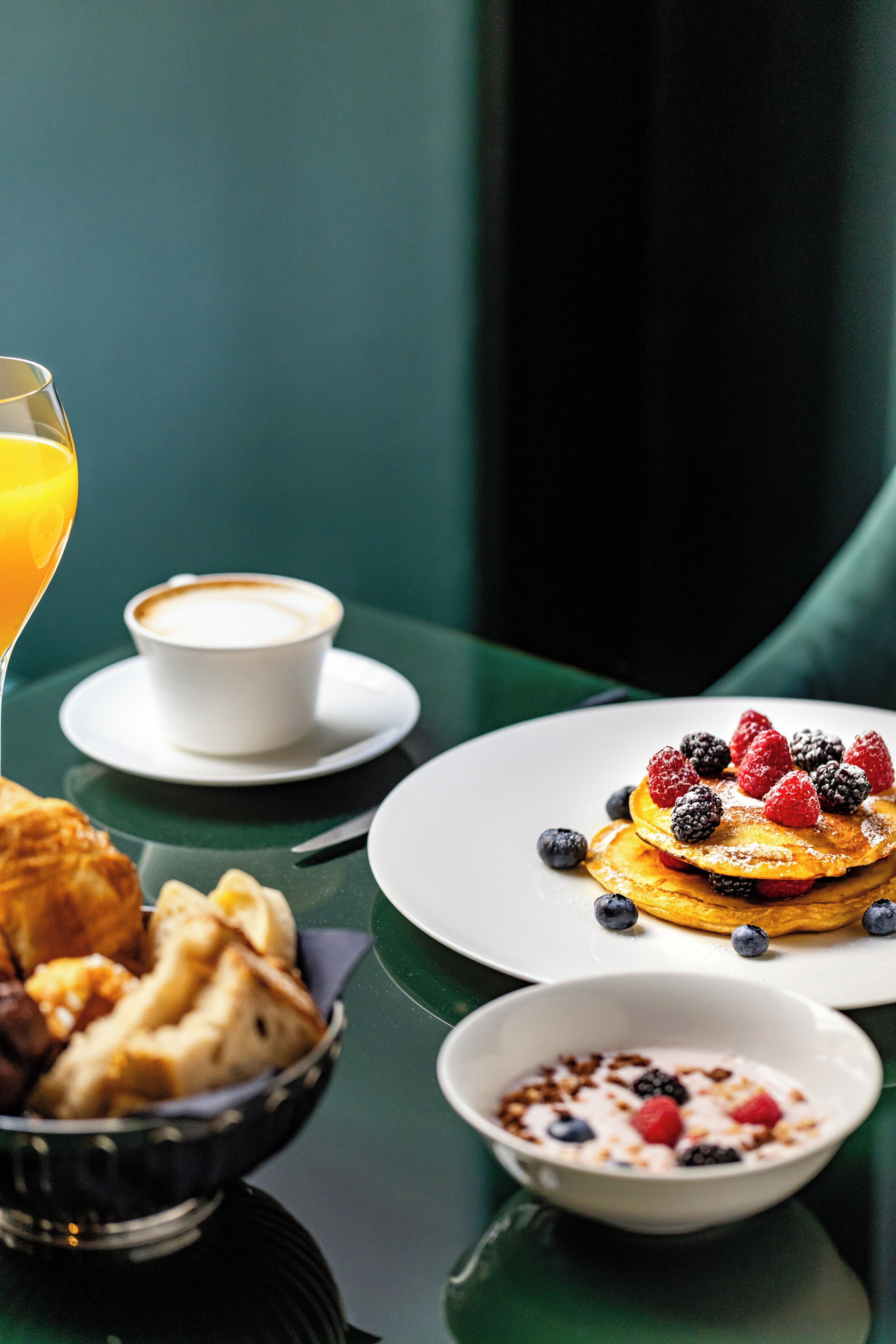 Ein Frühstückstisch mit Pfannkuchen, belegt mit Beeren, einem Glas Orangensaft, einer Tasse Kaffee und einer Schüssel Müsli.