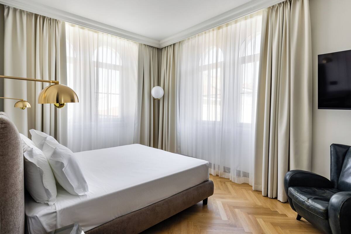 Una cama bien hecha con sábanas blancas, un sillón de cuero y un ventanal con cortinas traslúcidas.