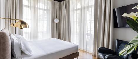 Une chambre à coucher moderne avec un lit, un fauteuil en cuir noir, un téléviseur fixé au mur et des rideaux légers.