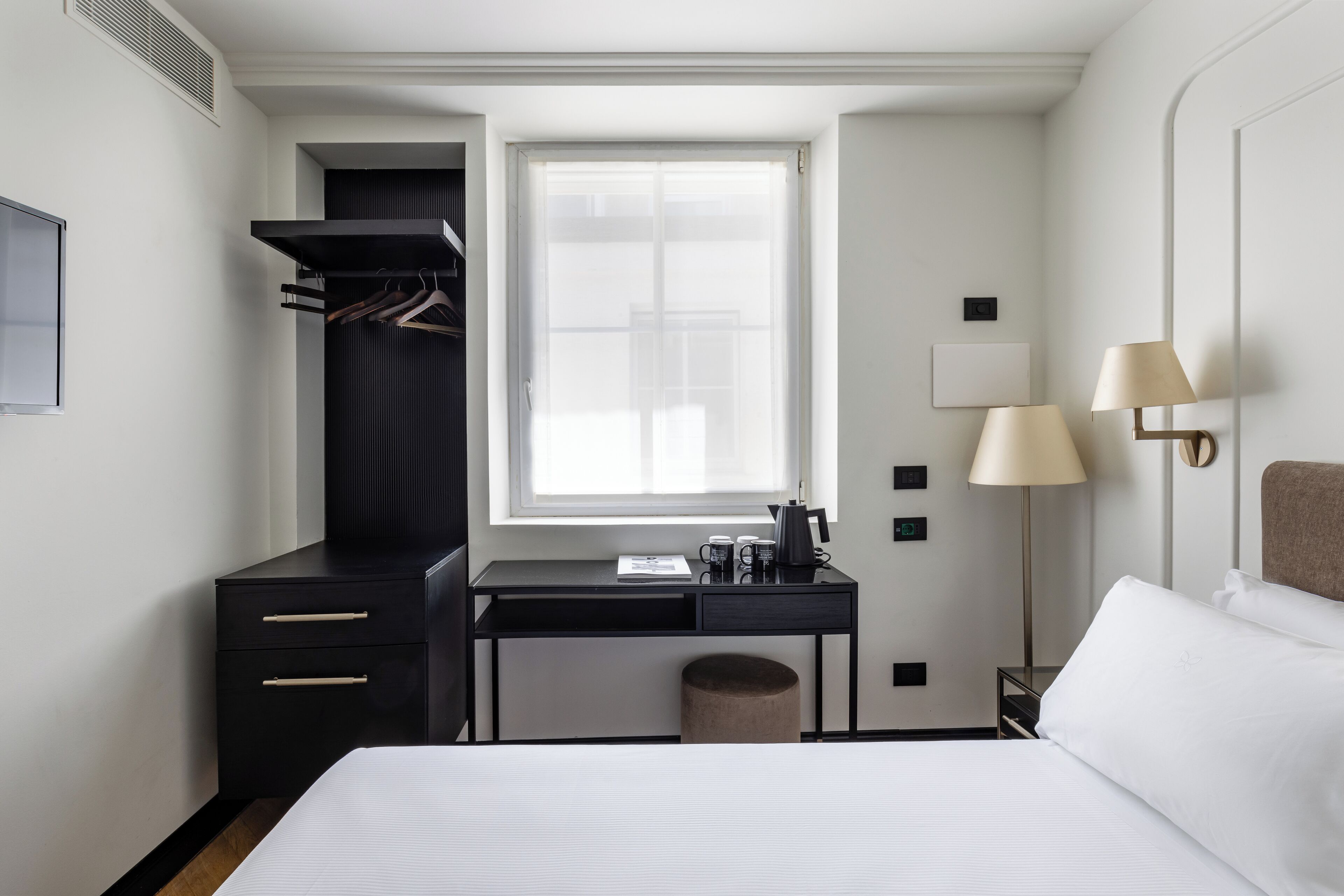 Une chambre d’hôtel comprenant un lit, un bureau avec une chaise, une armoire, une fenêtre et deux lampes de chevet.