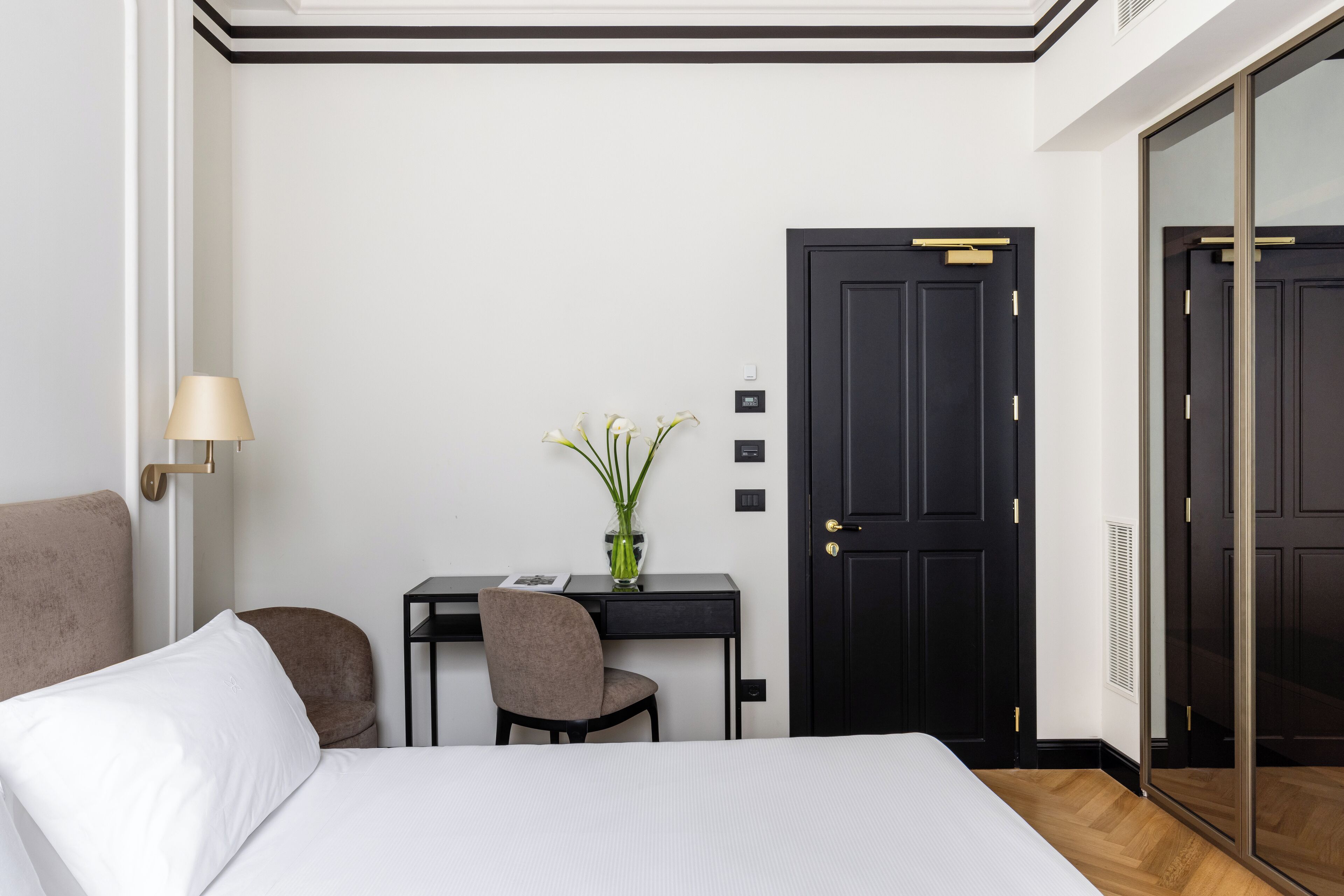 A hotel room with a bed, a desk with a chair, a vase with flowers, and a black door.