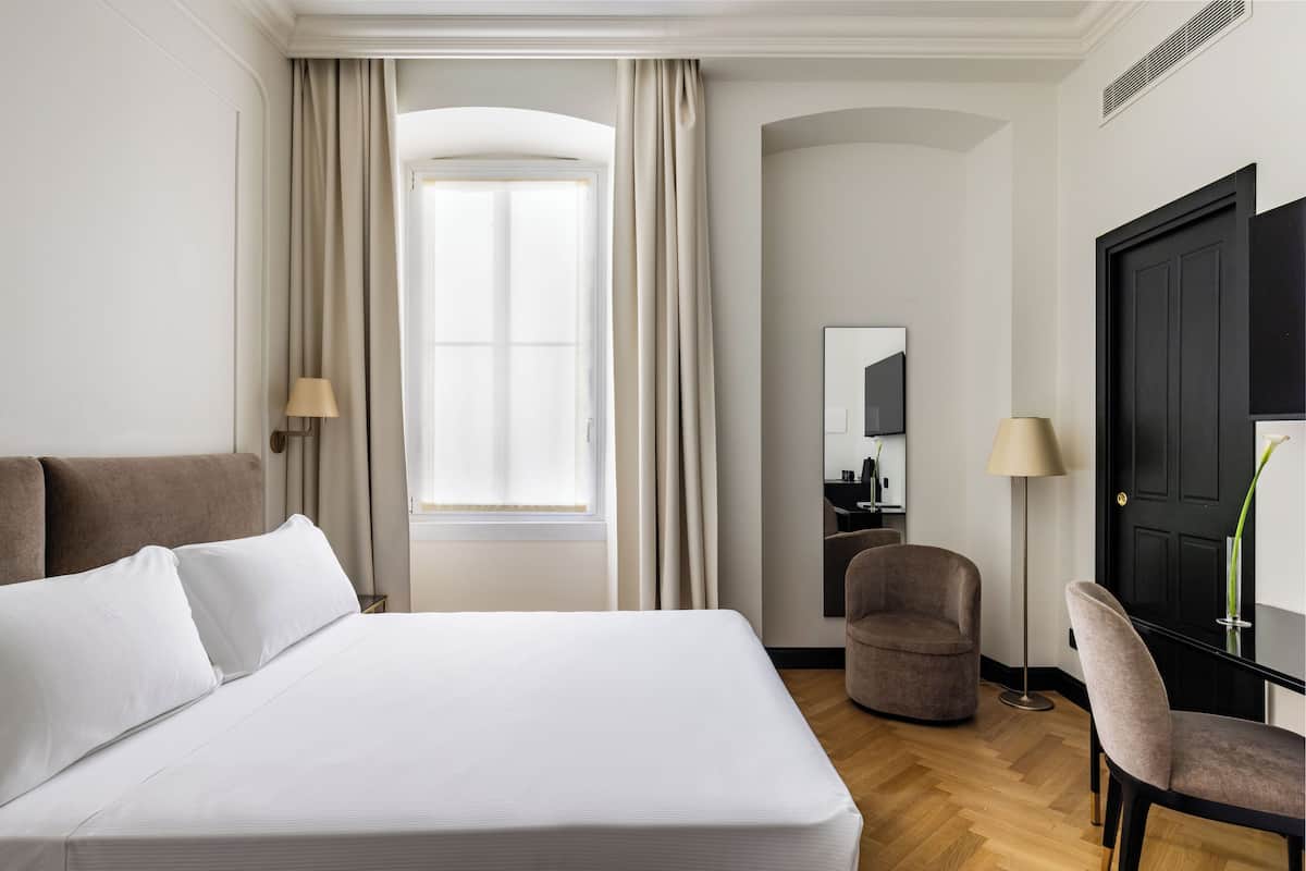 Una habitación de hotel con cama, una silla, un escritorio y un amplio ventanal con cortinas.