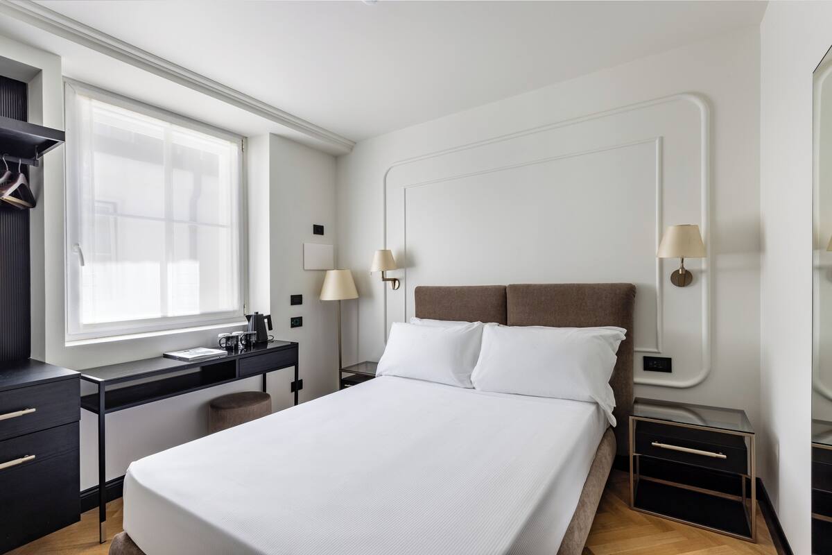 Una habitación de hotel moderna con una cama grande, un escritorio y una silla. Hay dos mesitas de noche con lámparas, una ventana con cortinas y un armario.
