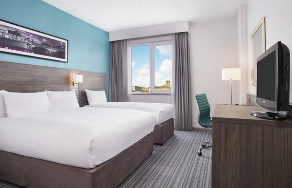 A hotel room with a large bed, a desk with a computer, a chair, and a window with curtains.