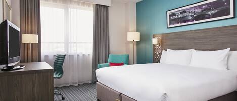 A hotel room with a large bed, a desk with a computer, a chair, a window with curtains, a wall-mounted lamp, and a framed picture.