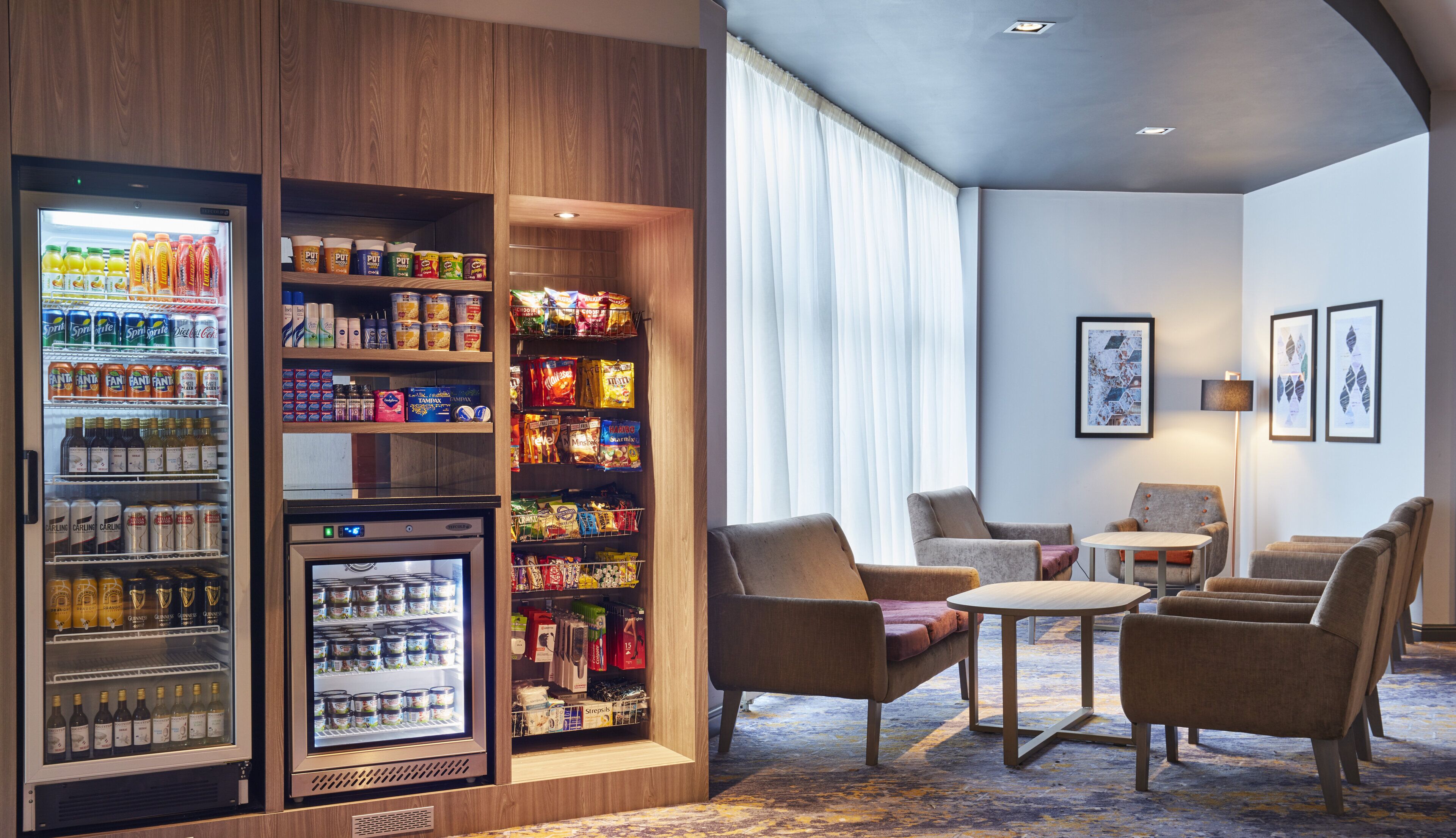A hotel lobby with a beverage fridge stocked with drinks, a snack bar, and seating area with chairs and a small table.