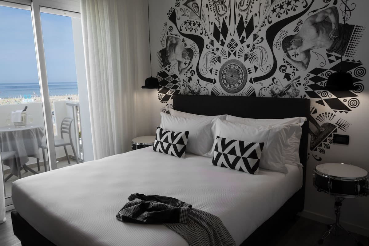 A hotel room with a large bed, a view of the beach, a patterned wall, and a drum set.