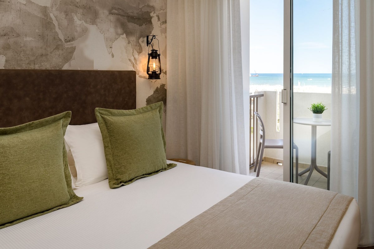 A hotel room with a large bed, green pillows, a world map on the wall, a lantern, and a balcony with a table and chair.