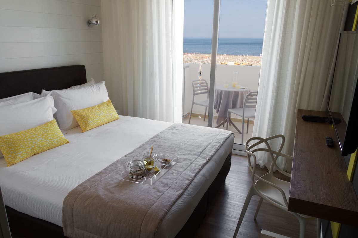 A hotel room with a bed, a small table with a tray of items, a chair, a desk, and a view of the beach.