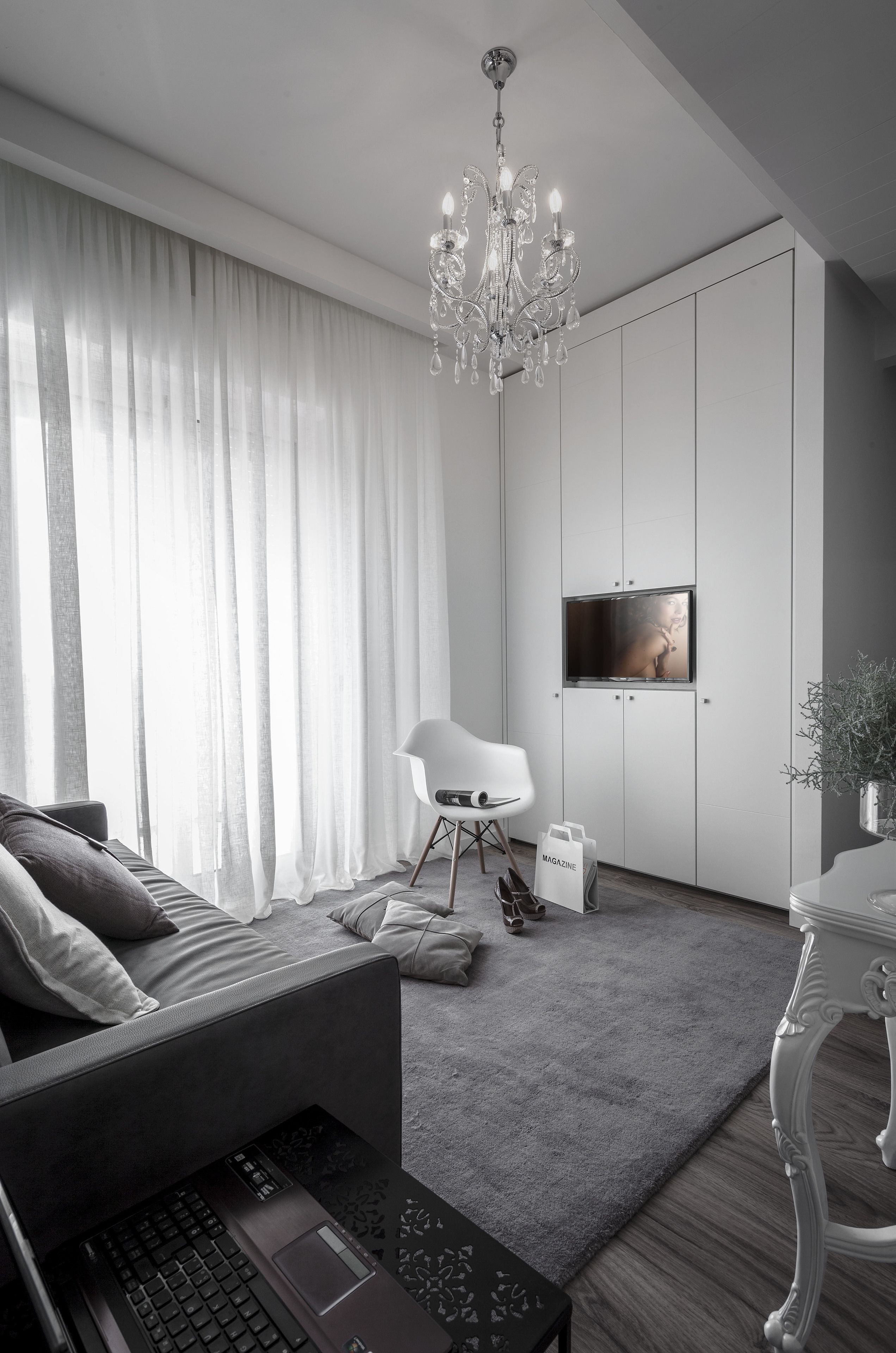 Una sala de estar moderna con un sofá gris, una computadora portátil sobre una mesa de centro, un sillón blanco, una araña y un televisor de pantalla plana montado en un mueble blanco.