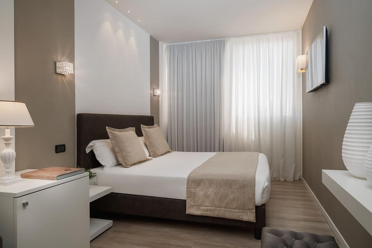 A modern hotel room with a large bed, a bedside table with a lamp, a mirror, and a view of a cityscape through sheer curtains.