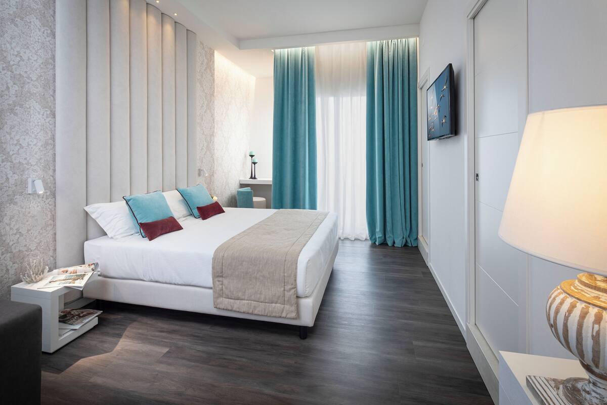 A modern hotel room with a large bed, a bedside table with a lamp, and a view of the outside through curtains.