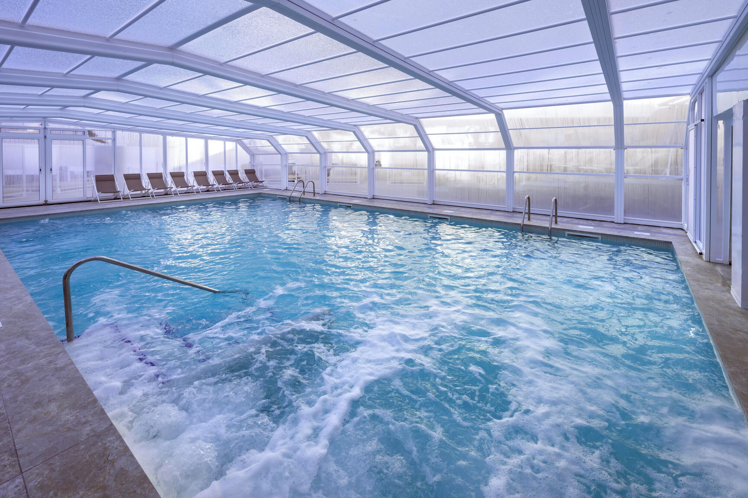 Une piscine intérieure avec des parois en verre transparent, un escalier et des chaises longues disposées le long du bord.