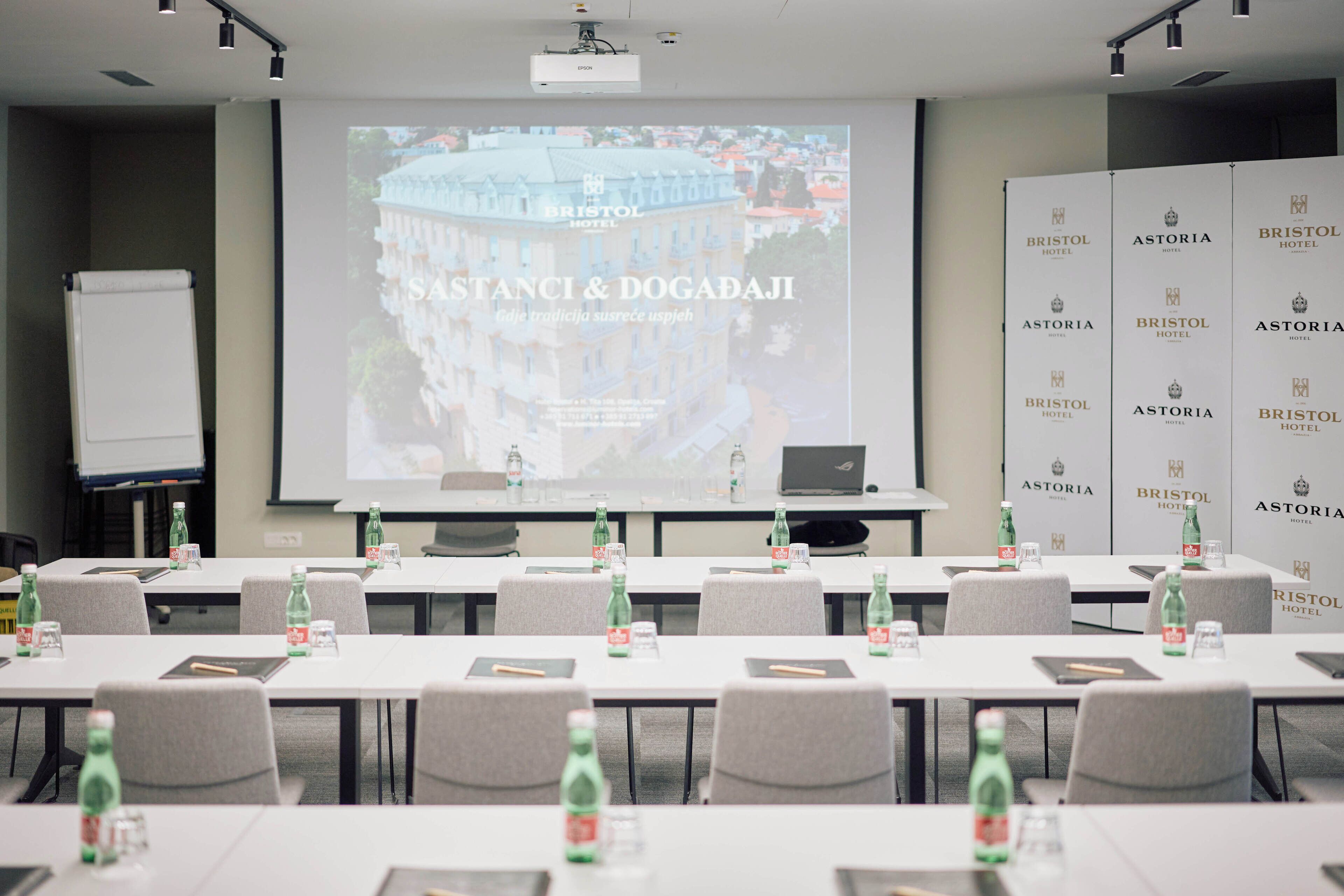 A conference room with a projection screen displaying a building and the text "Bristol Hotel." Tables are set with water bottles and notebooks.