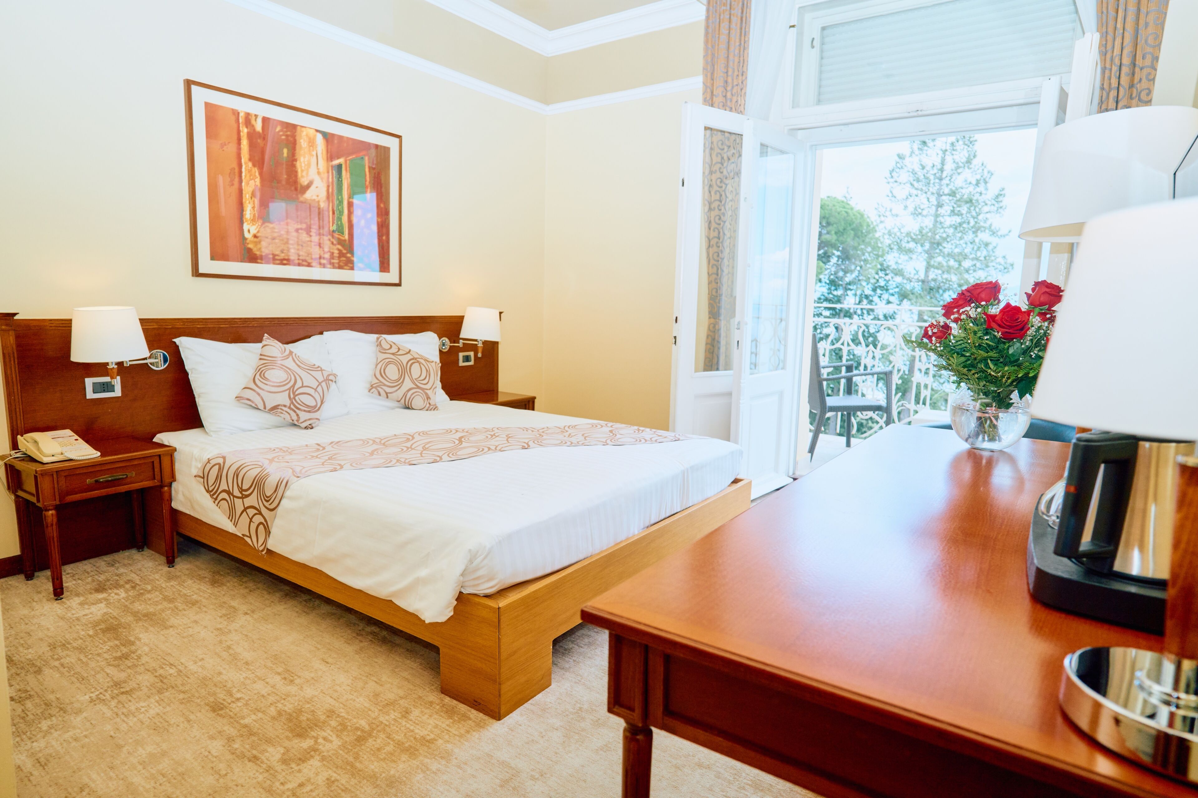 A hotel room with a bed, bedside tables, a coffee maker, a window with a view, and a painting on the wall.