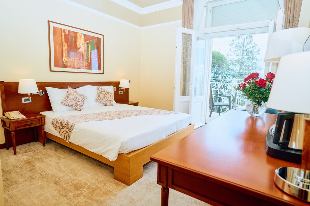 A hotel room with a bed, bedside tables, a coffee maker, a window with a view, and a painting on the wall.