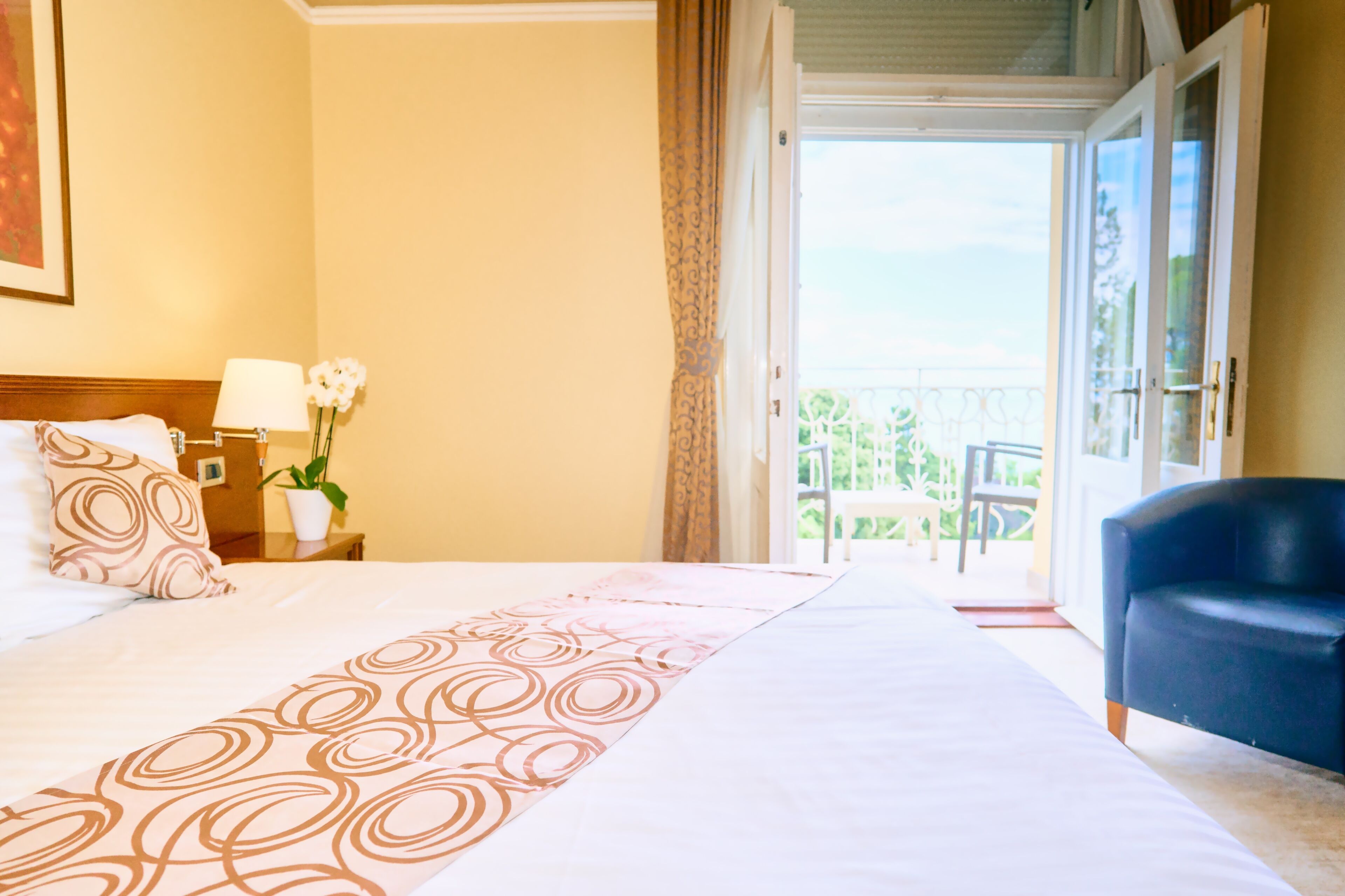 A hotel room with a bed, a chair, a nightstand with a lamp, and a view of the outdoors.