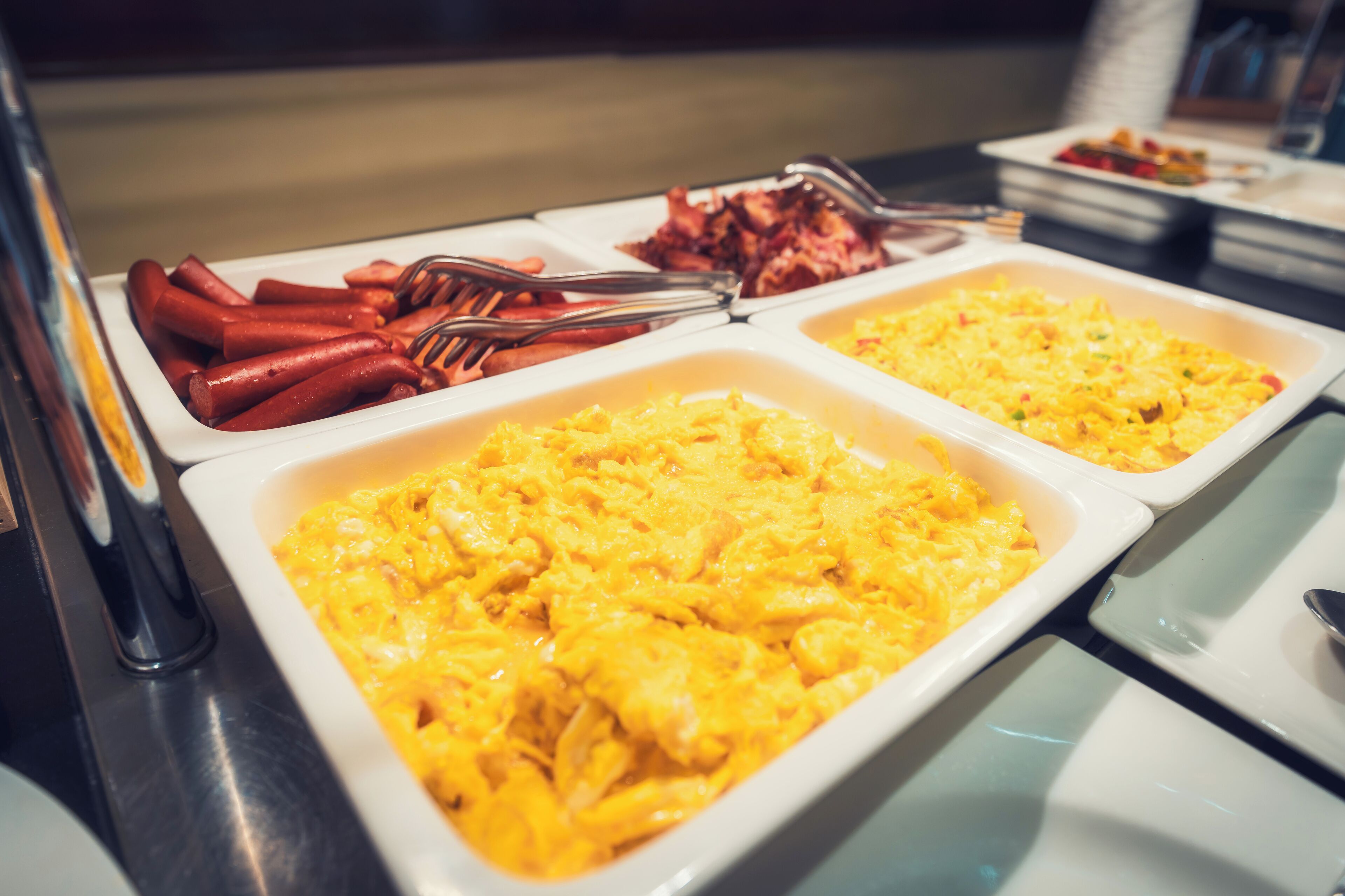 A buffet with various dishes including scrambled eggs, bacon, and hot dogs.