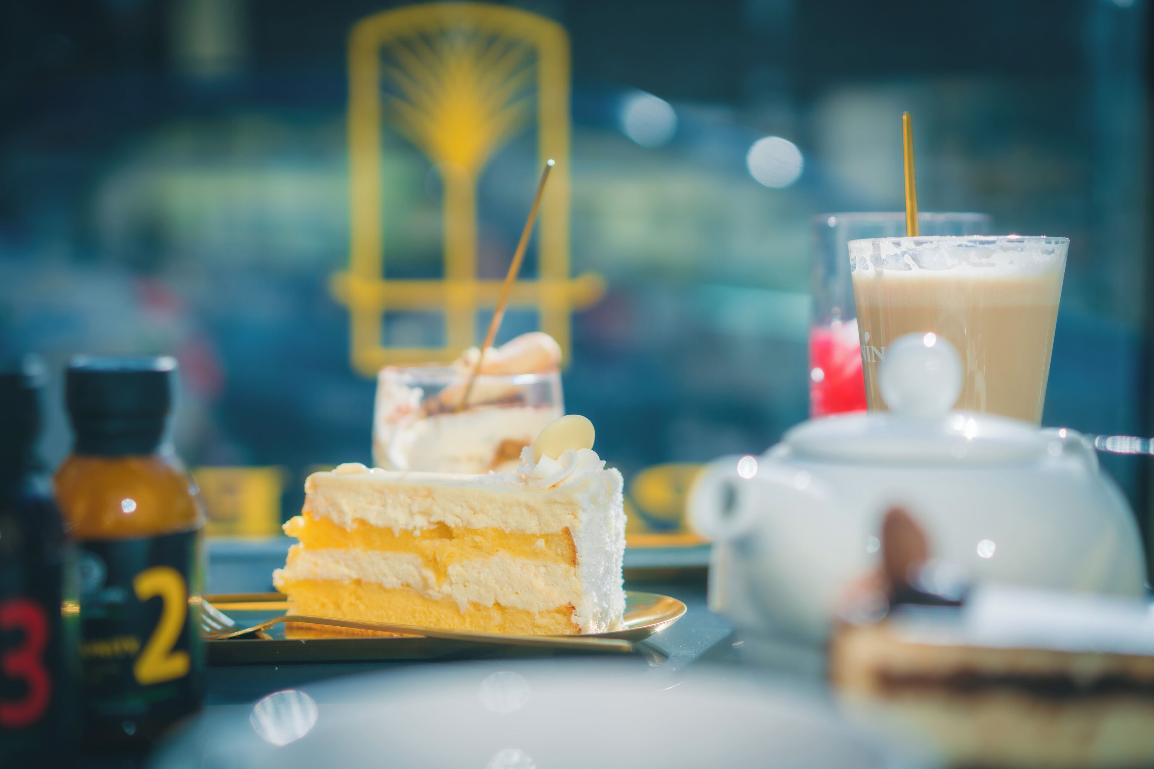 A dessert table with a slice of cake, a glass of iced coffee, and a teapot.