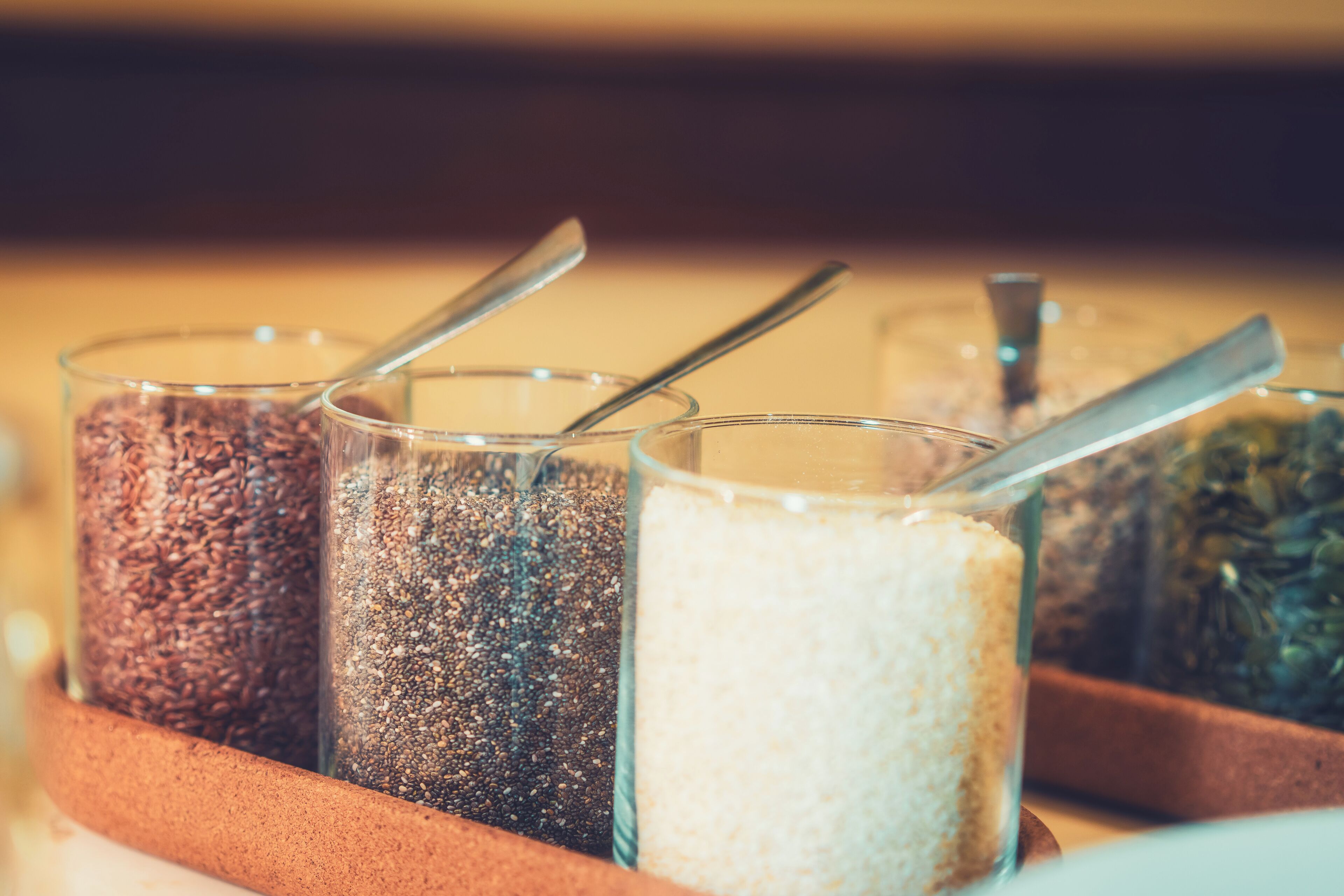 Four glass containers with spoons, each filled with different types of grains or seeds.
