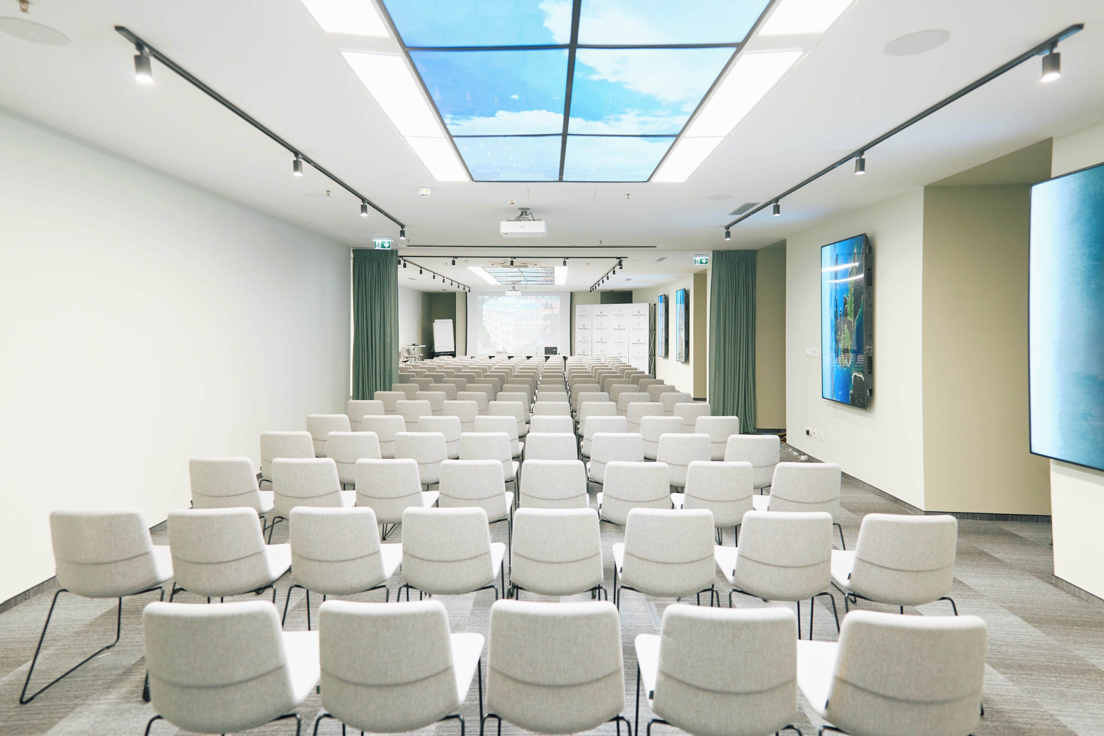 A conference room with rows of chairs, a large screen, and a ceiling with skylights.