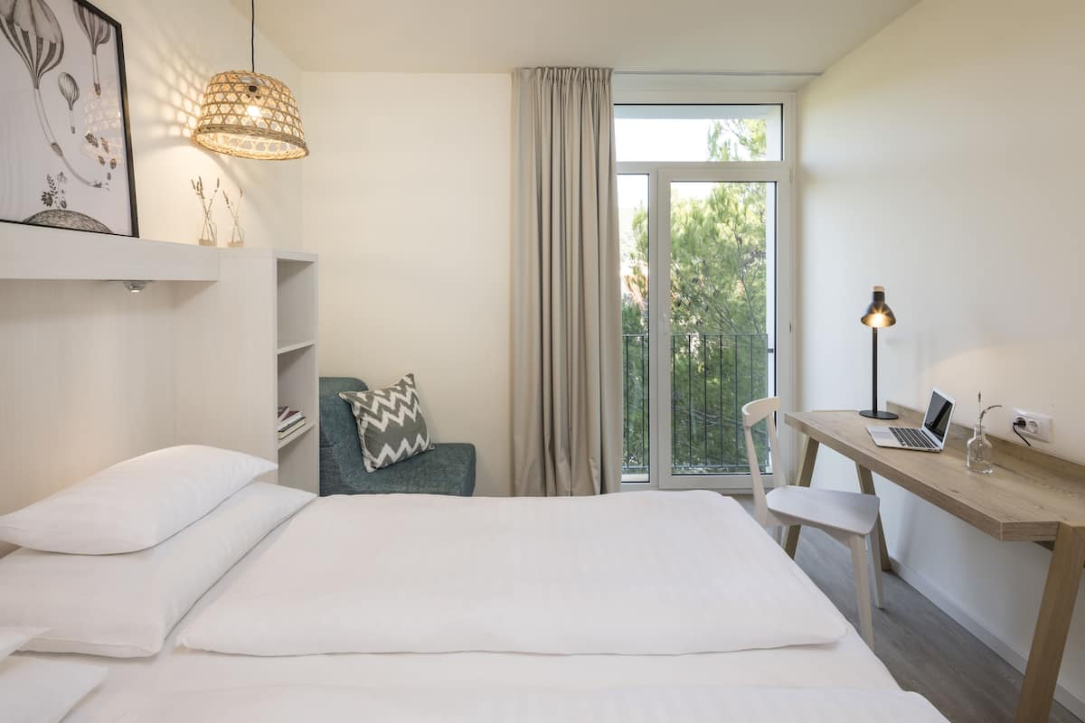 A modern hotel room with a bed, a desk with a laptop, a chair, a lamp, and a window with curtains.