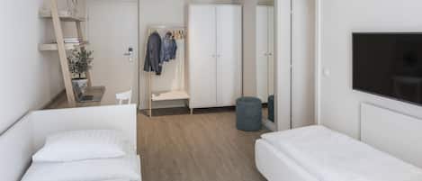 Un dormitorio moderno con dos camas individuales, un armario, un televisor y un pequeño rincón de descanso con una silla y una planta.