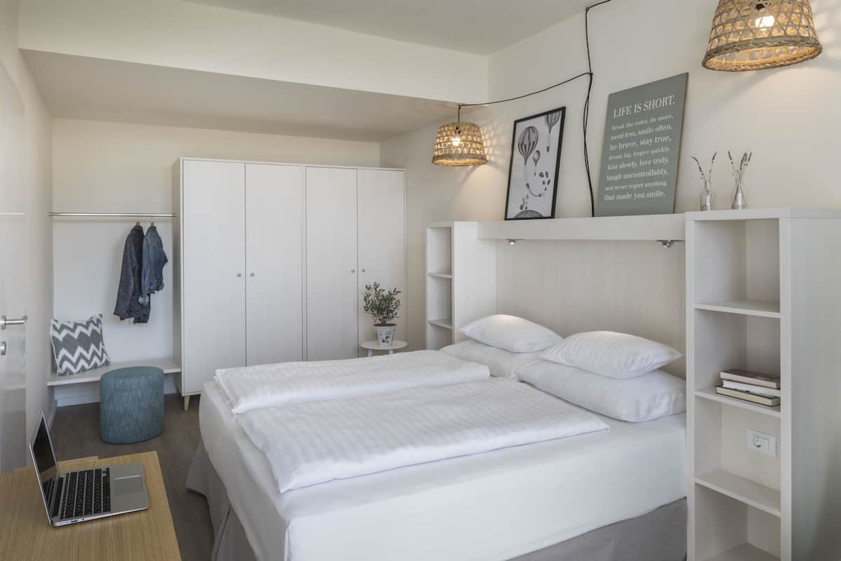A modern bedroom with a large bed, a wardrobe, a desk with a laptop, and a hanging light fixture.