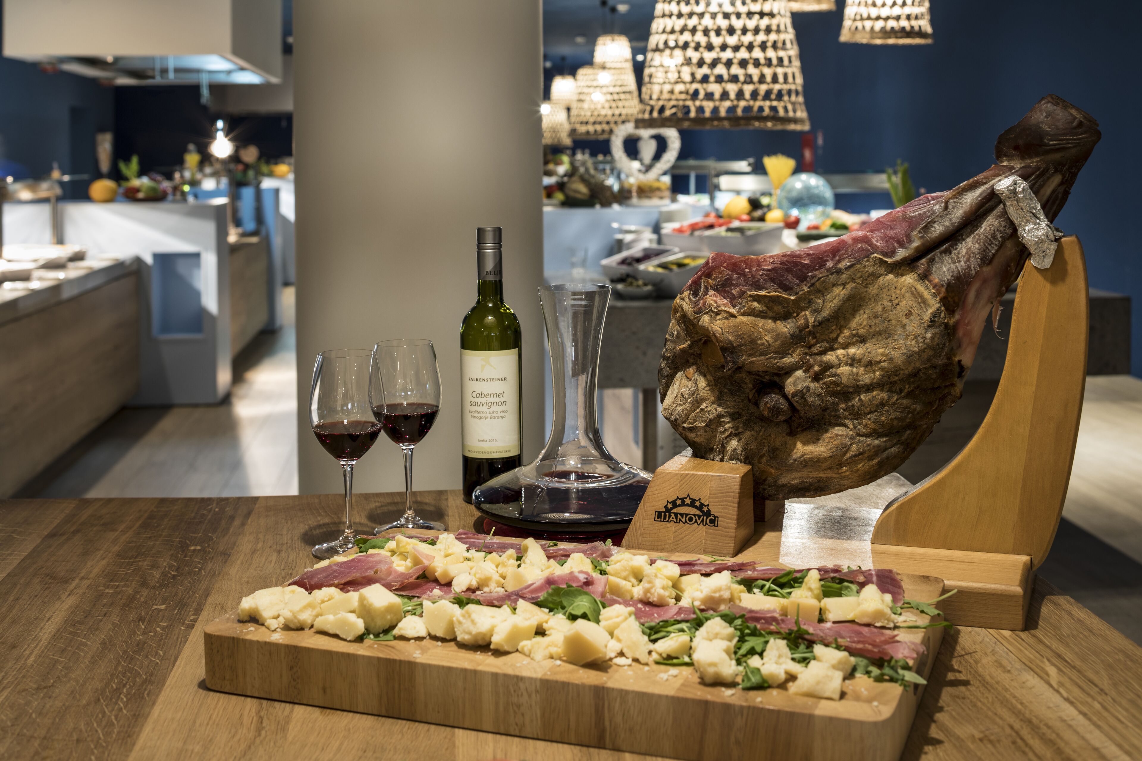 A wooden cutting board with cheese, meat, and greens, a bottle of wine, and a large piece of meat on a stand.