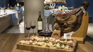A wooden cutting board with cheese, meat, and greens, a bottle of wine, and a large piece of meat on a stand.