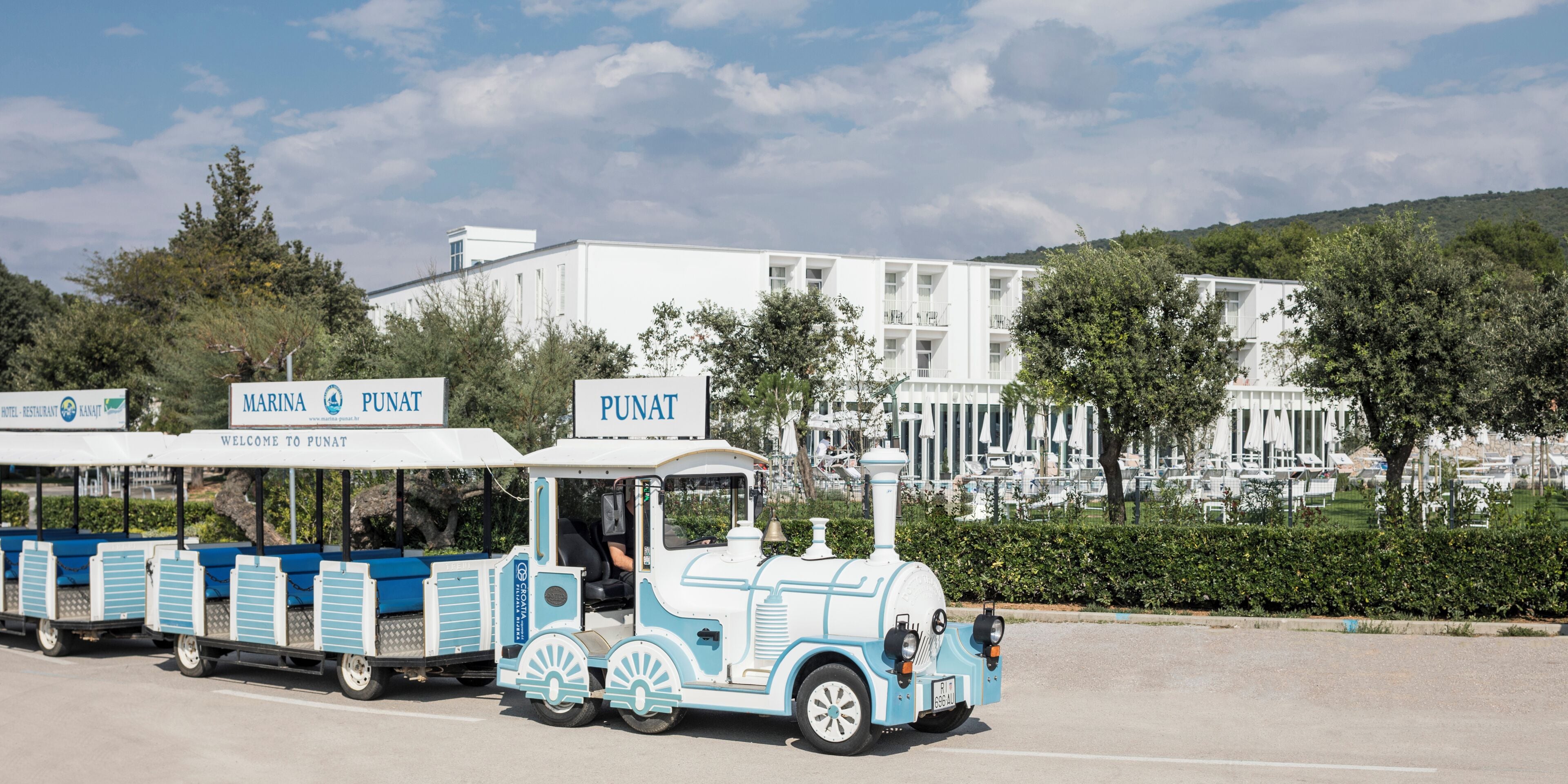 Un train bleu et blanc portant l’inscription « MARINA PUNAT », circulant sur une route avec un bâtiment blanc et des arbres en arrière-plan.