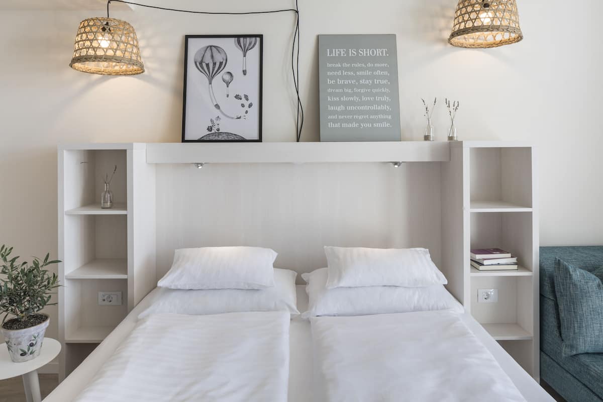A modern bedroom with two beds, a wall-mounted shelf with books, a framed quote, and a potted plant.