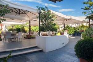 Zona patio esterna con tavoli e sedie sotto grandi ombrelloni, circondata da piante in vaso e un fioriere bianco.