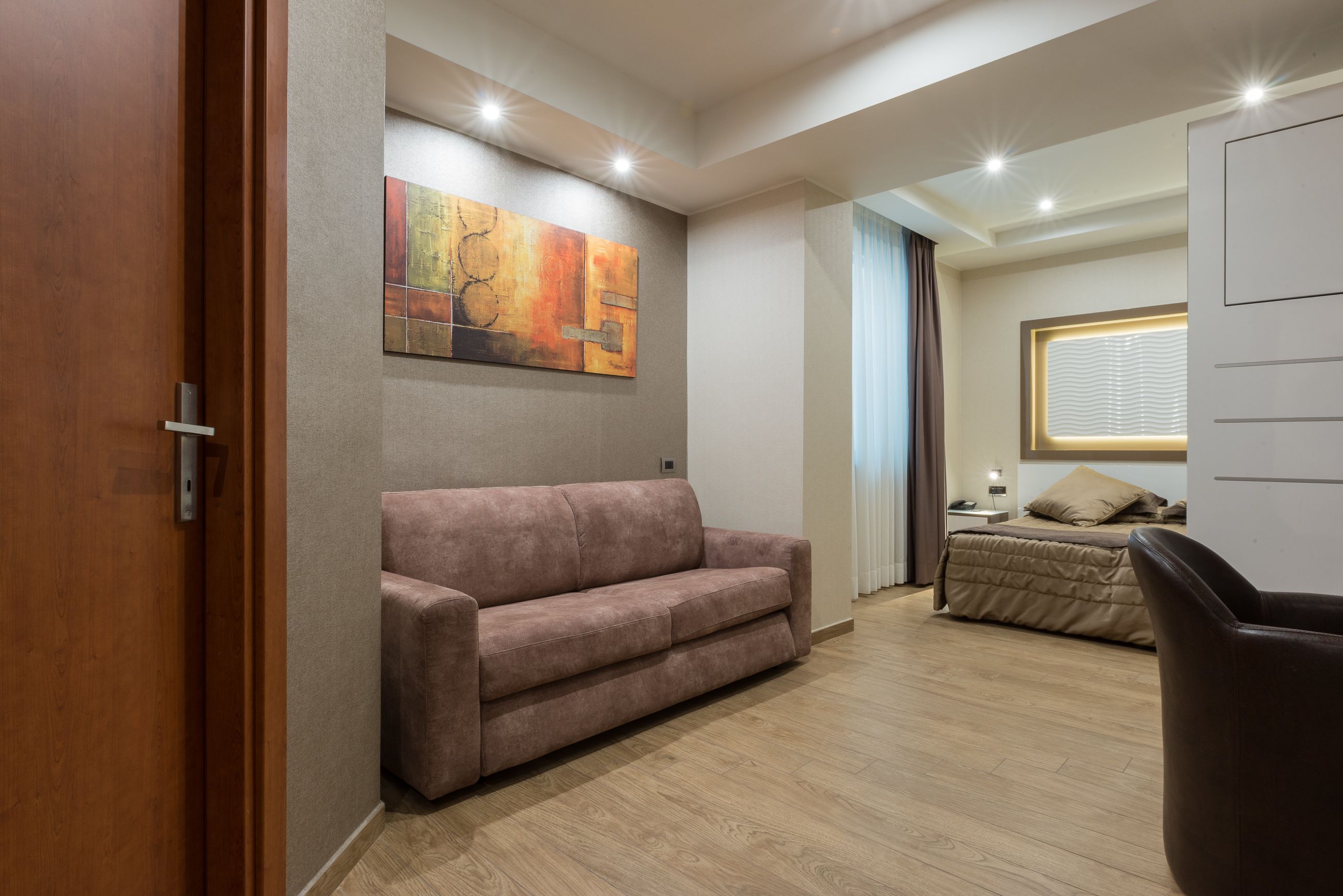 Quarto moderno com chão de madeira, um sofá marrom, cama com edredom bege e uma grande pintura abstrata na parede.