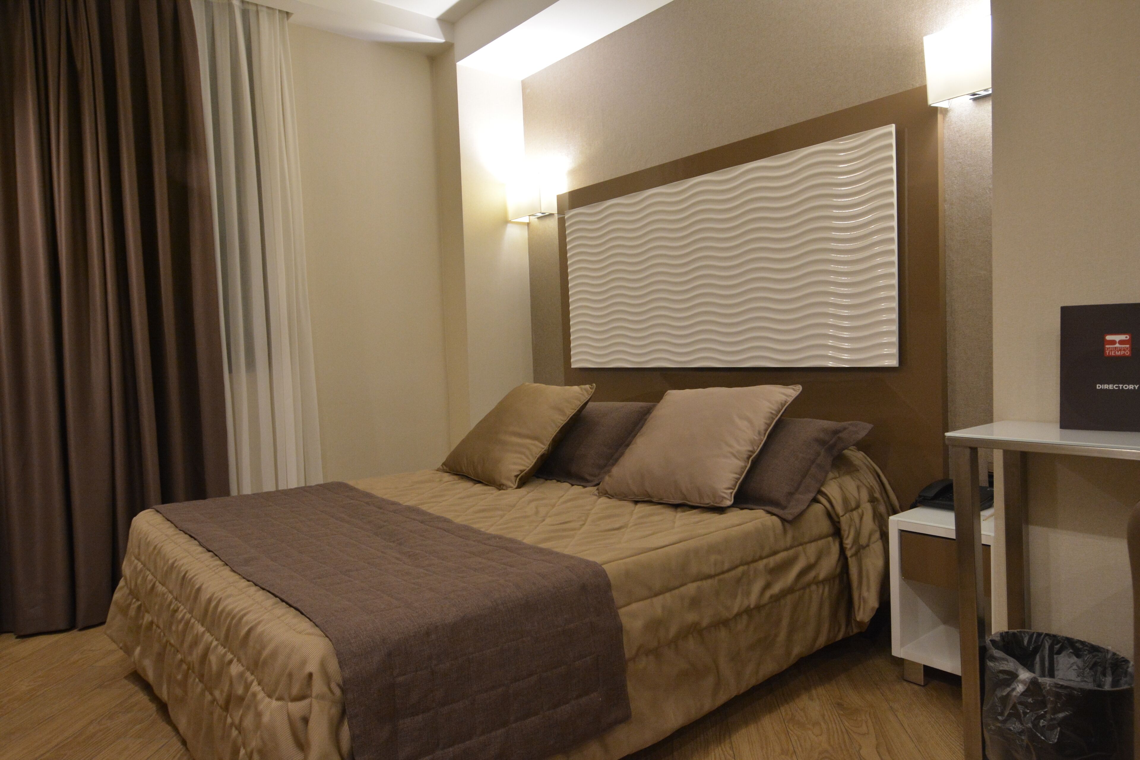 Una camera d'albergo con un letto, un comodino, un cestino e un lampadario a parete.