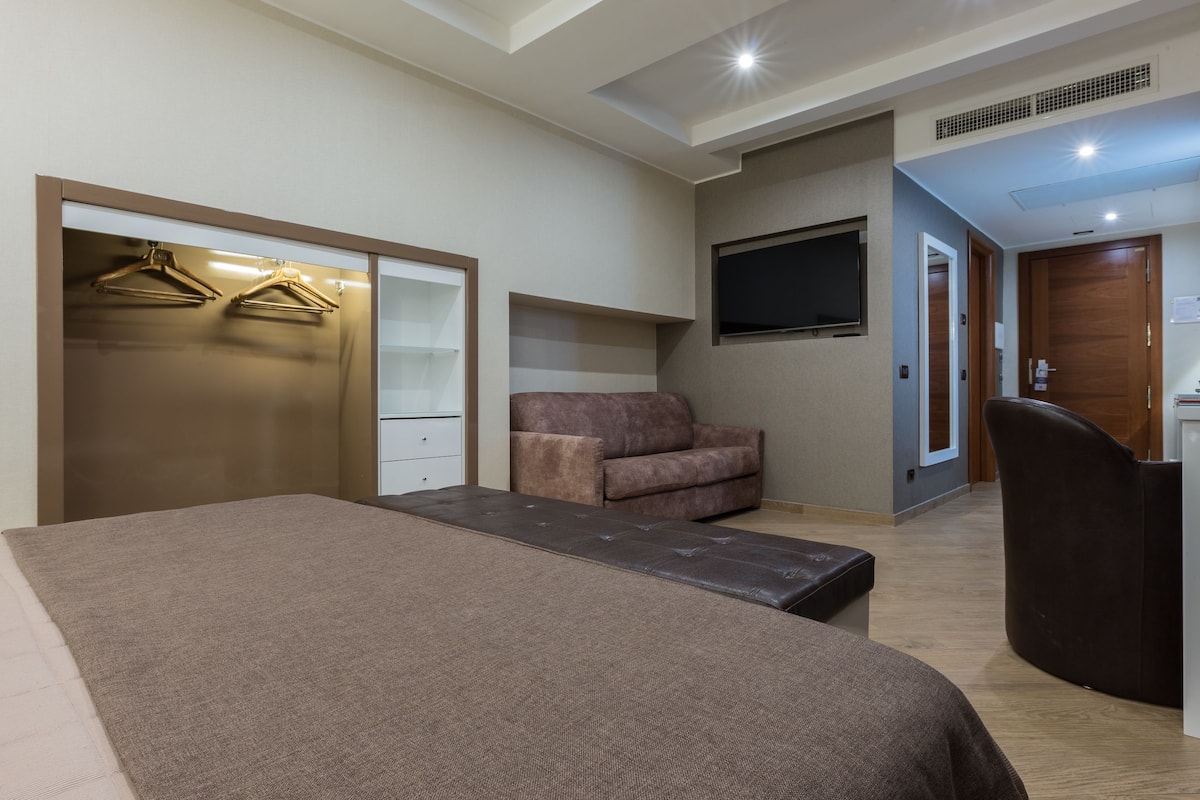 A modern hotel room with a bed, a seating area, a TV, and a wardrobe with hangers.
