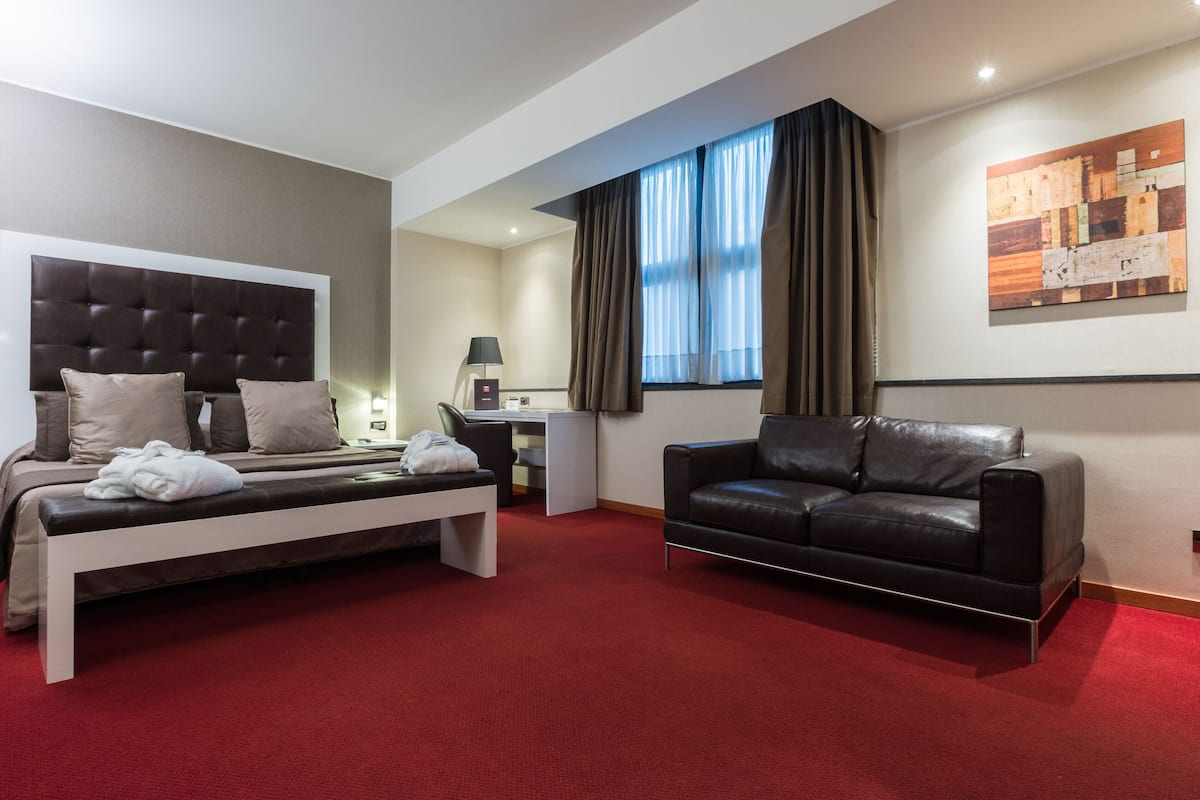 A hotel room with a bed, a desk, a black leather sofa, and a large window with curtains.