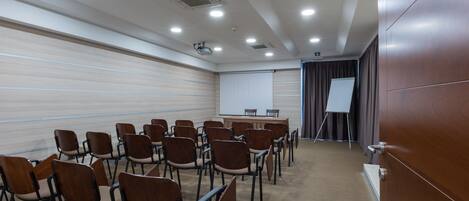 Un salón de conferencias con filas de sillas, una pantalla para proyector, una pizarra y una mesa de presentaciones.