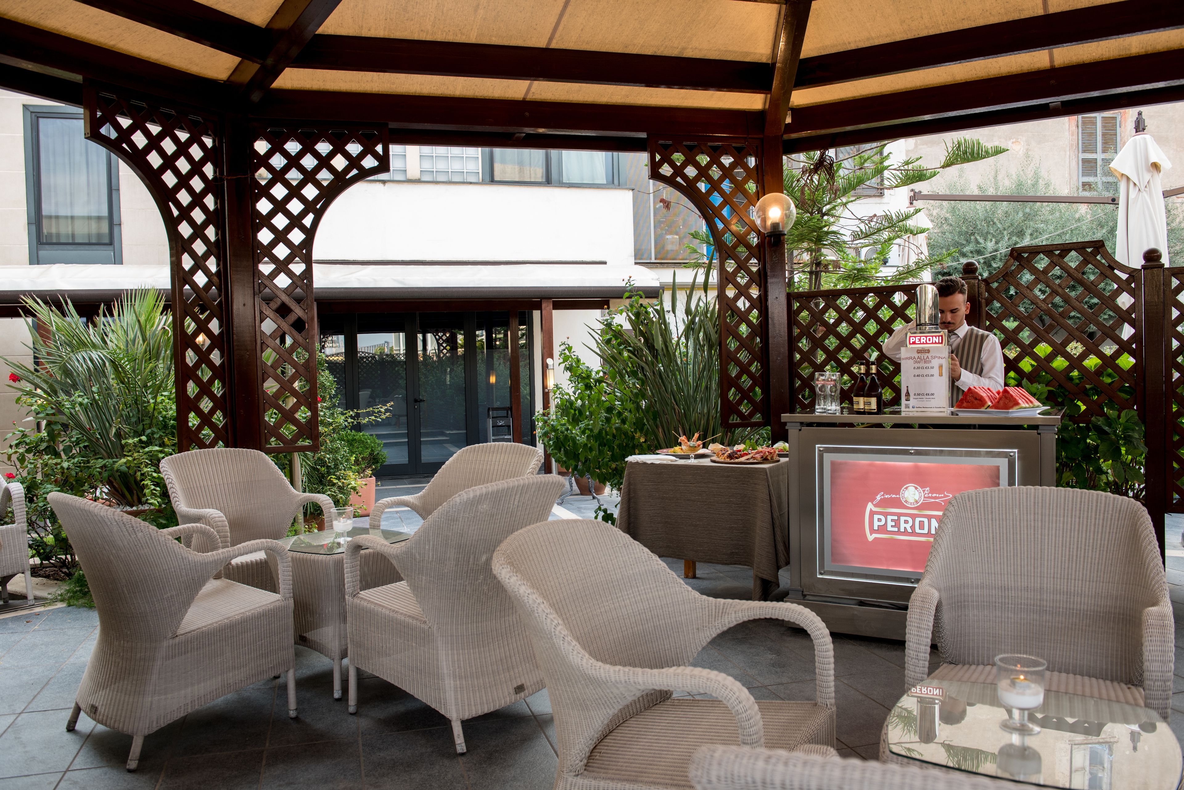 Un espace extérieur aménagé avec des chaises et une table en osier, un bar orné d’une enseigne Peroni et des plantes en pot.