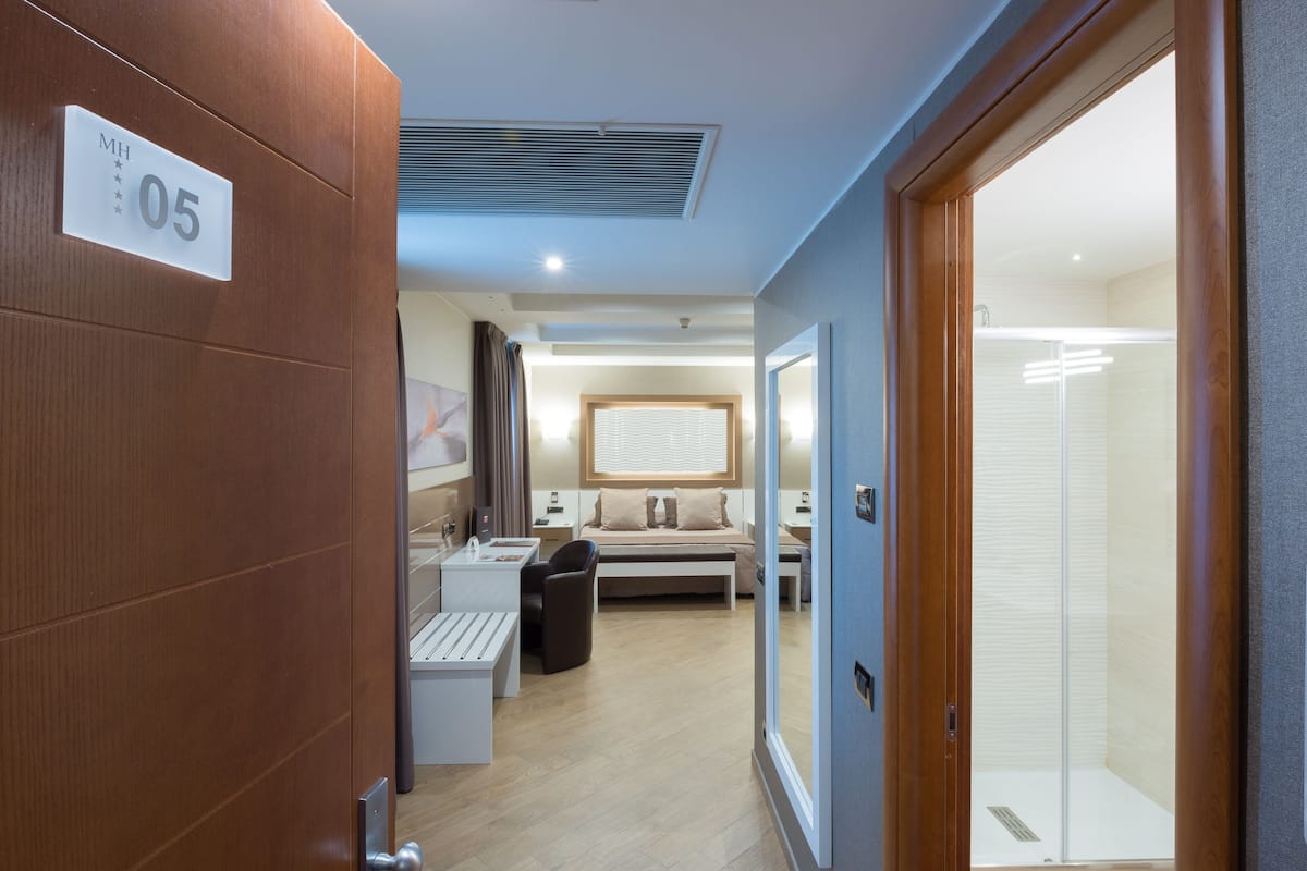 A hotel room with a bed, bedside table, and a small desk with a chair. There is a shower area visible through a glass door.