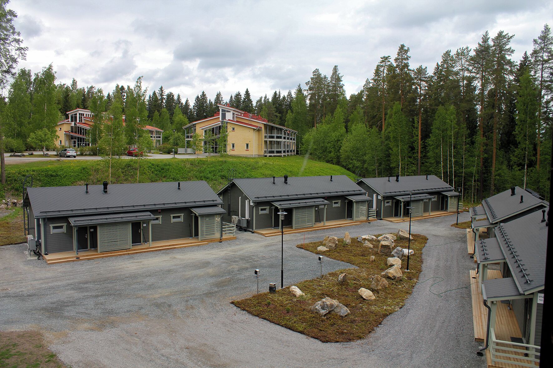 Une rangée de petits chalets modernes aux toits gris et aux bardages métalliques, situés dans une zone boisée, avec un grand bâtiment en arrière-plan.