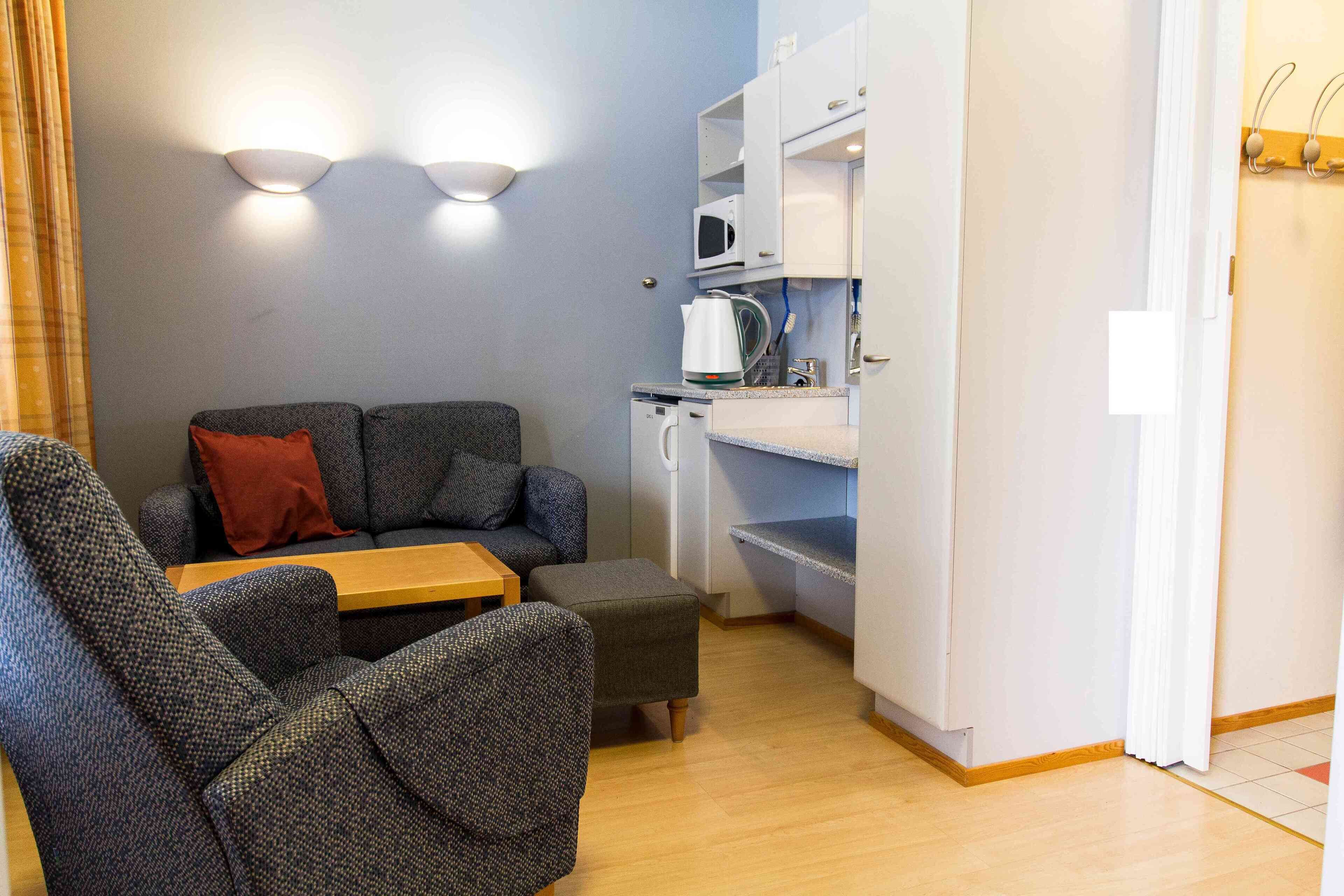 Un espace de vie compact comprenant un canapé, un pouf et une petite table. On peut voir un four à micro-ondes et une bouilloire sur le plan de travail.