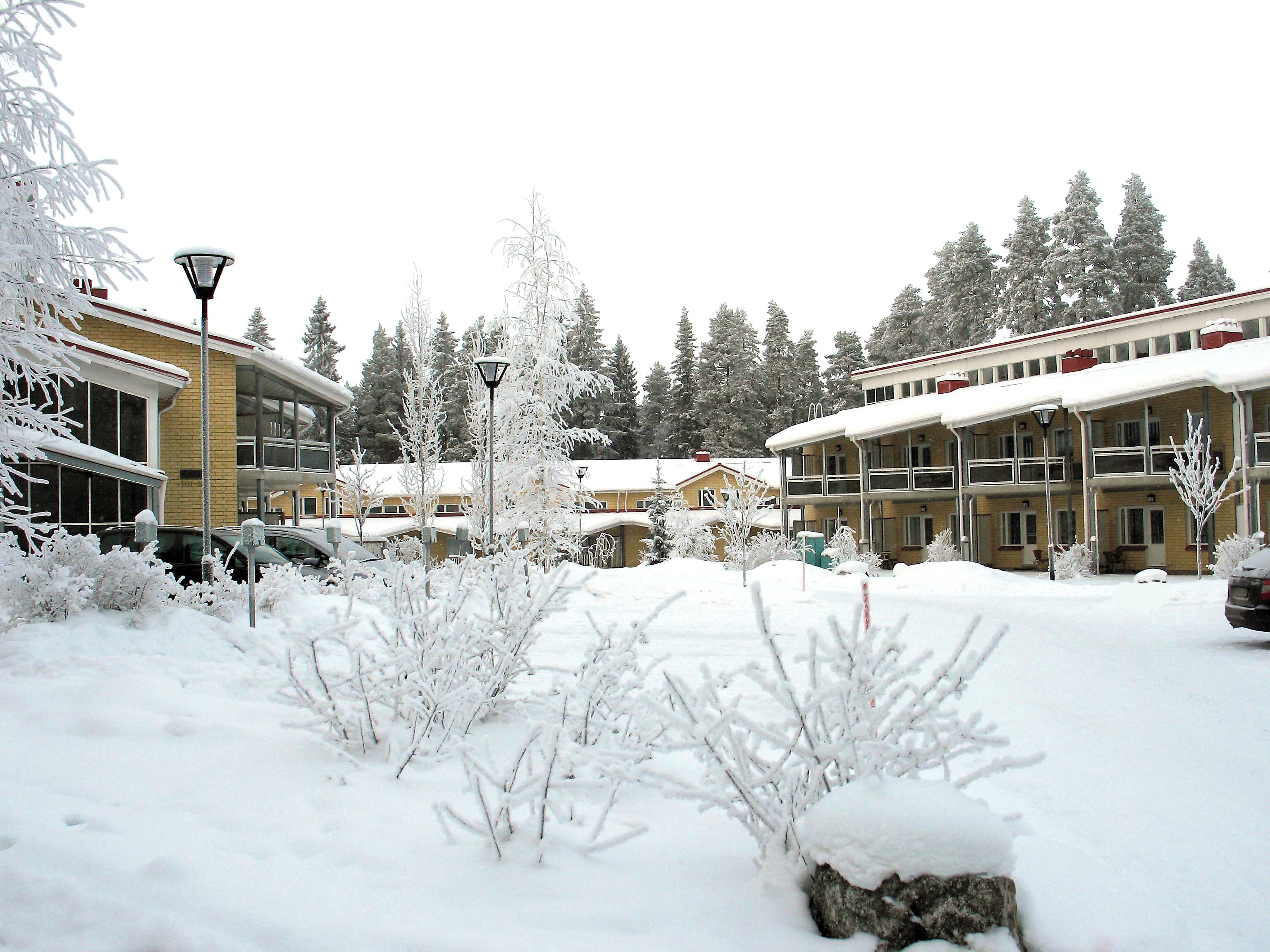 A snow-covered courtyard with buildings, including a two-story structure with balconies and a smaller building with a red roof.